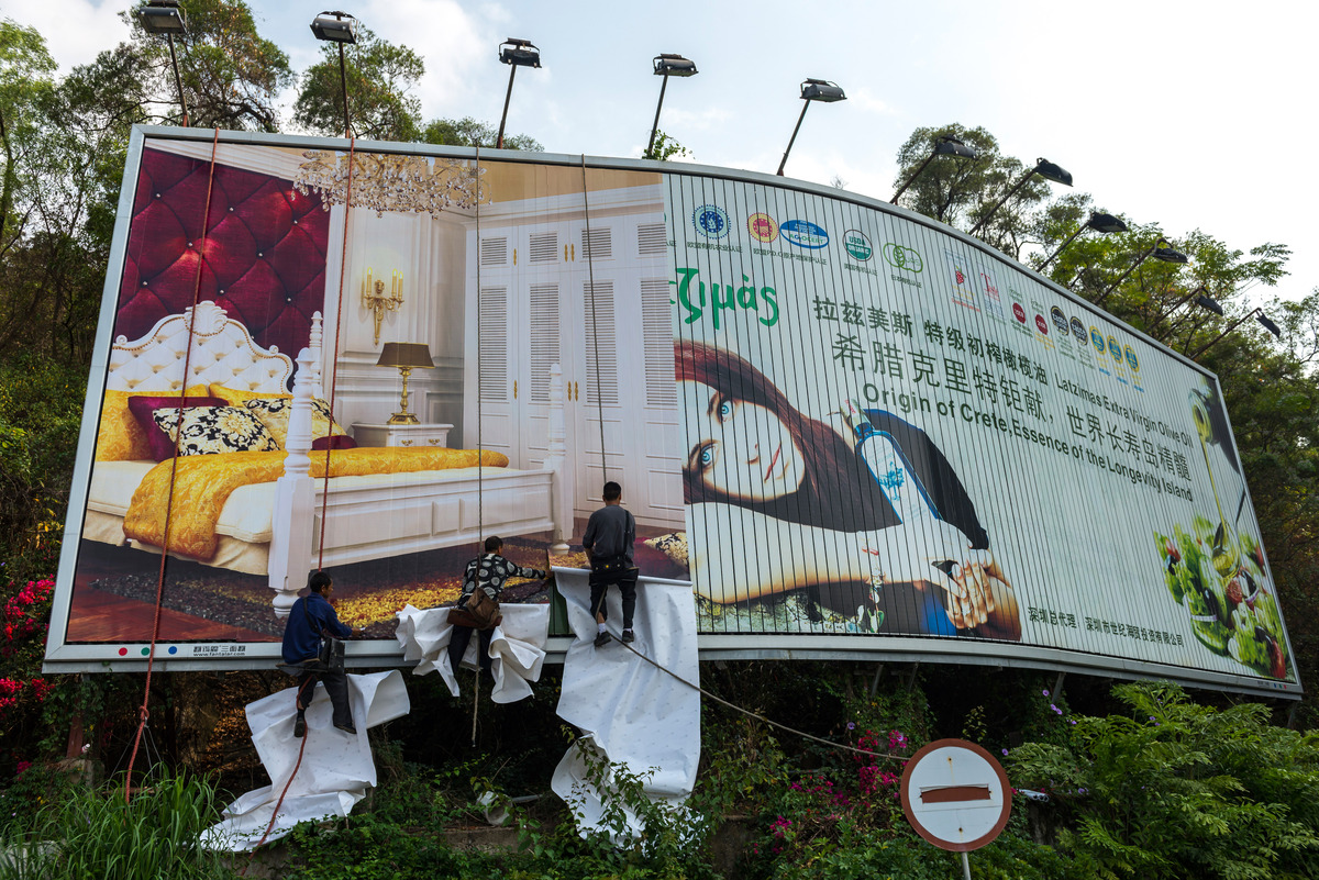 Trabalhadores instalando um grande outdoor publicitário com imagens de produtos e ambientes sofisticados, evidenciando como a repetição visual em peças de mídia exterior contribui para o efeito da mera exposição, ampliando o reconhecimento de marca e fortalecendo a comunicação visual e estratégica no espaço urbano.